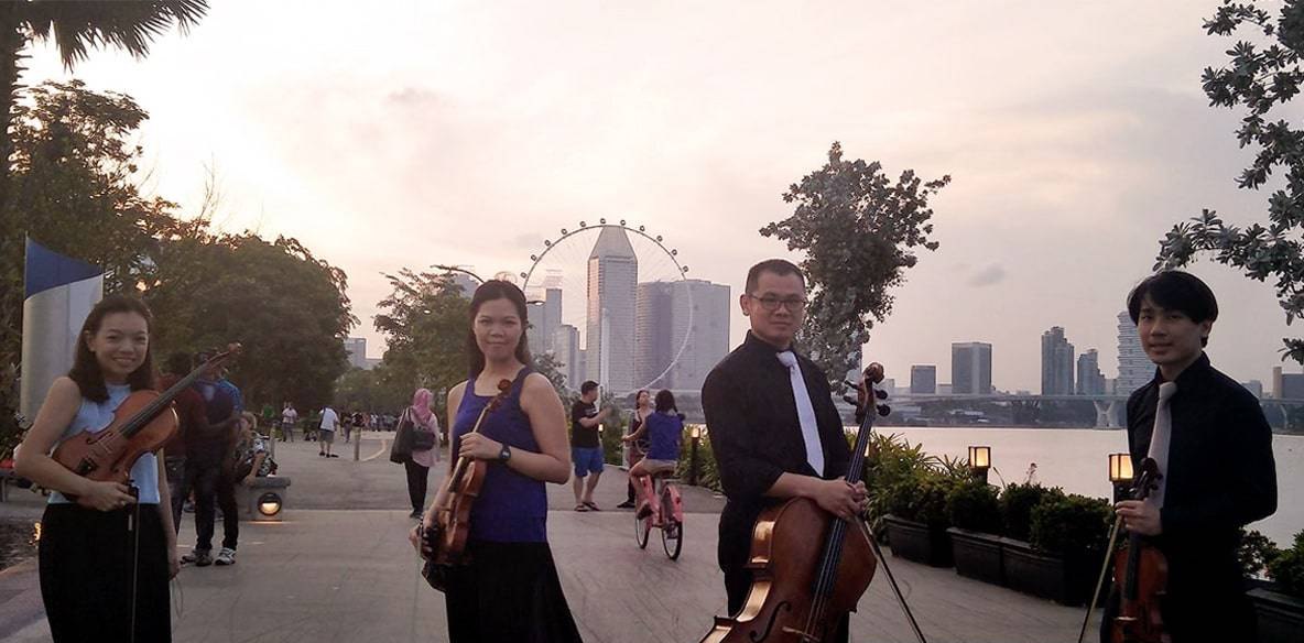 The GraceNotes string quartet standing by the water at Marina Bay at dusk, with the Singapore skyline behind them